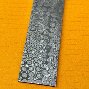 May include: Close-up of a rectangular piece of steel with a unique pattern. The steel has a dark gray color with a complex, swirling design of concentric circles and lines. The background is a solid, bright yellow.