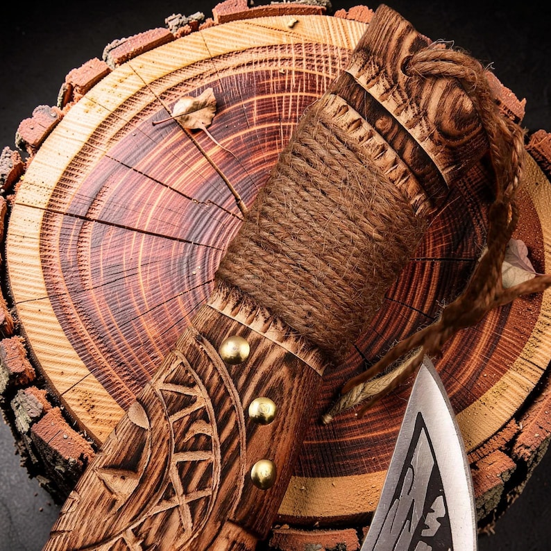 K&ouml;nnte beinhalten: Ein Handbeil mit einem dunkelbraunen Holzgriff, der geschnitzte geometrische Muster, drei Messingnieten und eine dicke Juteseilwicklung aufweist. Eine geflochtene Juteschnur ist am Knauf befestigt und eine scharfe Metallklinge ist sichtbar.