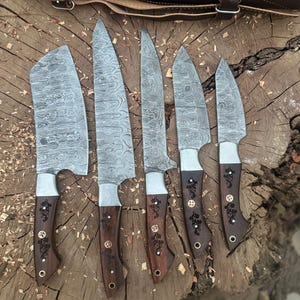May include: A set of six Damascus steel knives with wooden handles. The blades have a unique patterned design. The handles are dark brown with decorative metal accents. A brown leather knife roll is in the background.