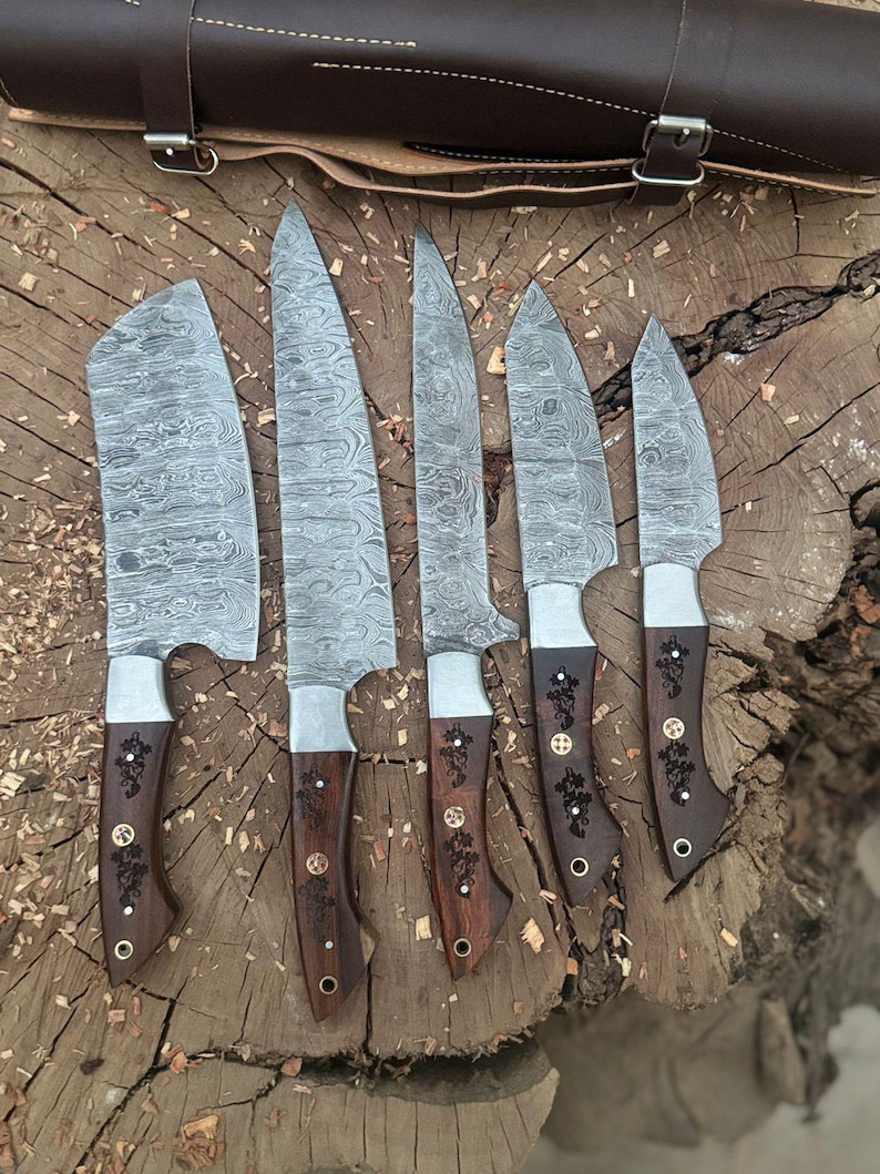 May include: A set of six Damascus steel knives with wooden handles. The blades have a unique patterned design. The handles are dark brown with decorative metal accents. A brown leather knife roll is in the background.