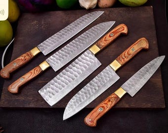 May include: A set of four kitchen knives with hammered metal blades featuring a visible layered pattern. Each knife has a wooden handle with a distinct grain, a brass-colored bolster, and a decorative rivet. The blades vary in shape and size.