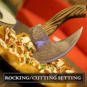 K&ouml;nnte beinhalten: Ein geschwungener Pizzaschneider aus Metall mit einem Holzgriff mit dunkler Maserung. Die Klinge ist mit einem Tannenzapfen, einem stilisierten Tierkopf und Runentext graviert. Der Text ROCKING/CUTTING SETTING steht unten im Bild.