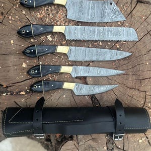May include: A set of five Damascus steel kitchen knives with black handles and brass bolsters, displayed on a wooden surface. The knives vary in size and shape, including a cleaver, bread knife, and paring knife. A black leather knife roll is included.