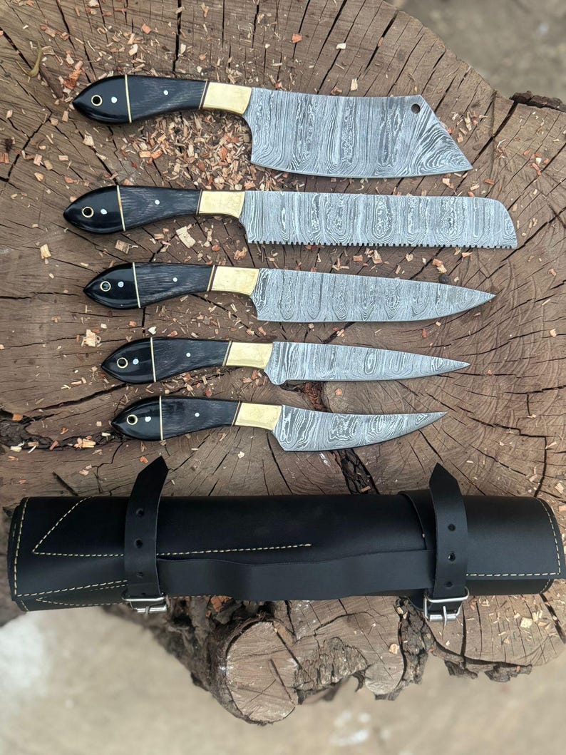 May include: A set of five Damascus steel kitchen knives with black handles and brass bolsters, displayed on a wooden surface. The knives vary in size and shape, including a cleaver, bread knife, and paring knife. A black leather knife roll is included.
