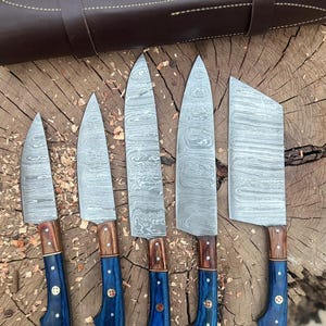 May include: A set of six Damascus steel kitchen knives with wooden and blue handles. The blades have a unique patterned design. The knives are arranged on a wooden surface, with a brown leather sheath in the background.