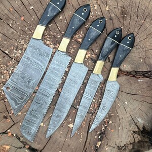 May include: A set of six Damascus steel knives with black handles, brass bolsters, and decorative rivets. The blades have a unique, wavy pattern. The knives are arranged on a wooden surface, showcasing their craftsmanship and design.