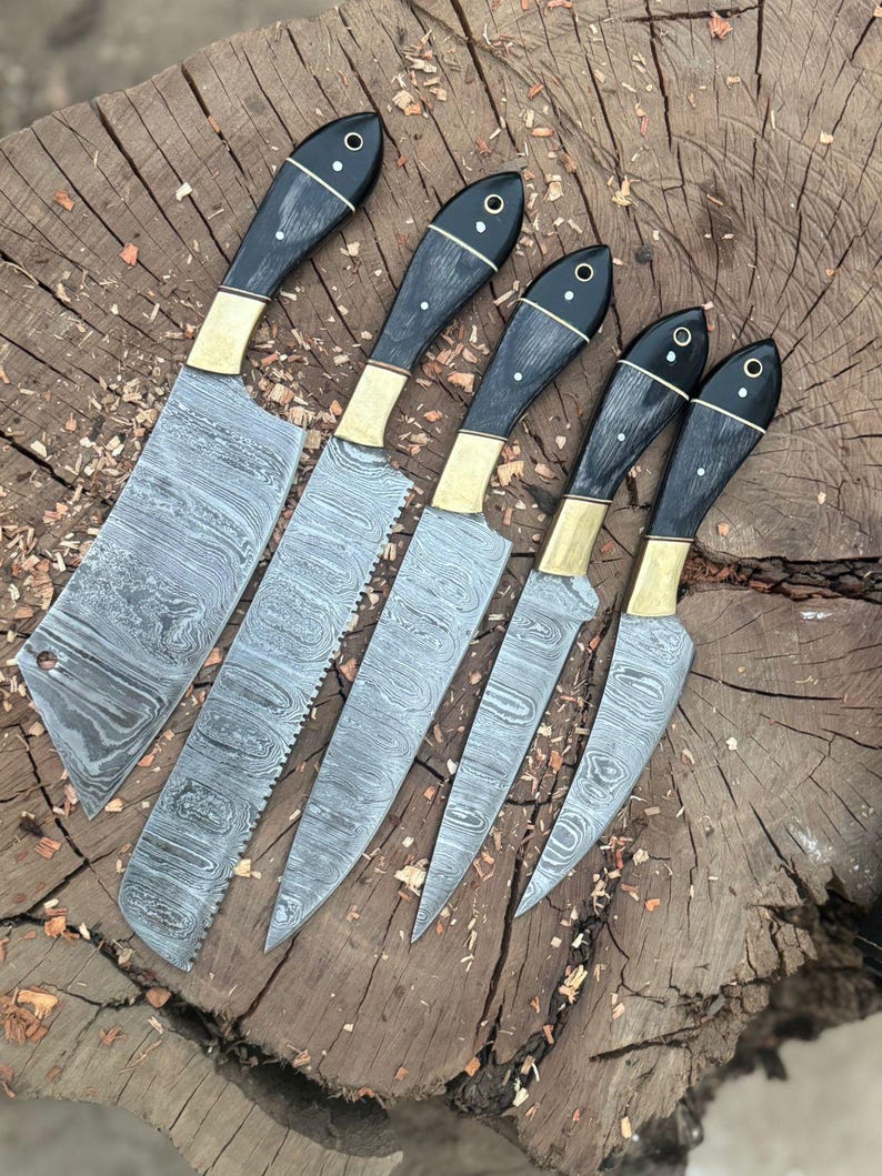 May include: A set of six Damascus steel knives with black handles, brass bolsters, and decorative rivets. The blades have a unique, wavy pattern. The knives are arranged on a wooden surface, showcasing their craftsmanship and design.