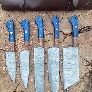 May include: A set of five kitchen knives with blue handles and patterned blades, displayed with a brown leather carrying case. The knives have wooden accents and are arranged on a wooden surface.