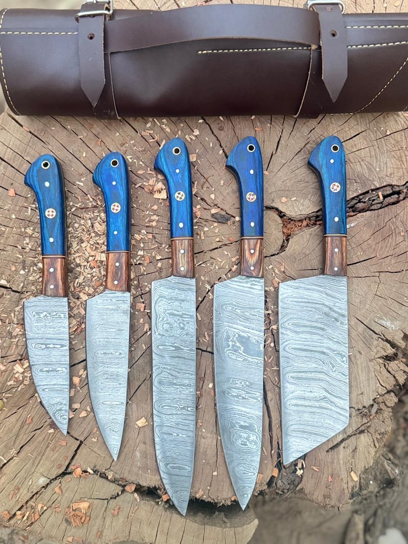 May include: A set of five kitchen knives with blue handles and patterned blades, displayed with a brown leather carrying case. The knives have wooden accents and are arranged on a wooden surface.
