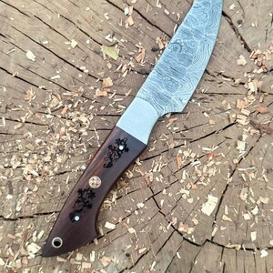 May include: A handcrafted knife with a patterned steel blade and a dark wood handle. The handle features decorative carvings and inlays. The knife is shown on a wooden surface with wood shavings.