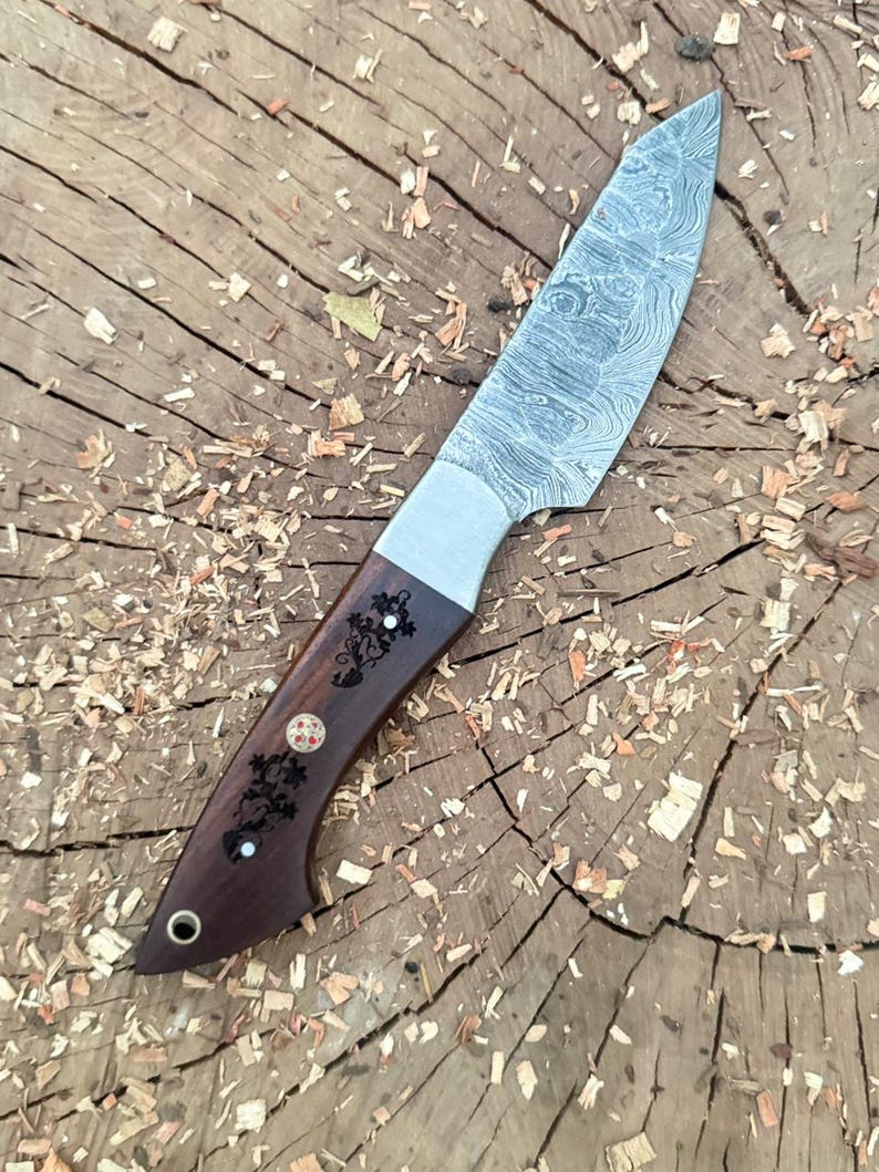 May include: A handcrafted knife with a patterned steel blade and a dark wood handle. The handle features decorative carvings and inlays. The knife is shown on a wooden surface with wood shavings.