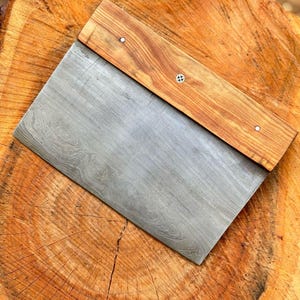 May include: A handmade, rectangular metal and wood scraper. The blade is a patterned gray, and the handle is a light brown wood, attached with small silver rivets. The scraper rests on a textured, brown wooden surface.