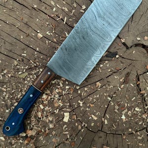 May include: A large, Damascus steel cleaver knife with a wooden and blue handle. The blade has a unique pattern and is set against a rustic wooden background. The handle is a combination of dark wood and blue, with decorative rivets.