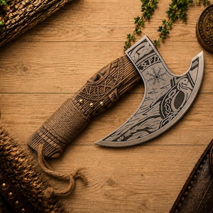 K&ouml;nnte beinhalten: Eine Handaxt mit einer geschwungenen Metallklinge, die mit nordisch inspirierten Mustern und einem Vegvisir-Symbol graviert ist. Der Griff besteht aus geschnitztem Holz mit komplexen Knotenmustern und ist mit einer Naturfaserkordel umwickelt.