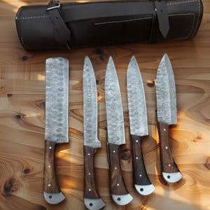 May include: A set of five forged steel knives with wooden handles and a leather carrying case. The blades have a textured finish, and the handles are dark brown with silver rivets. The leather case is dark brown with a buckle closure.
