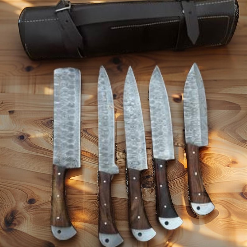 May include: A set of five forged steel knives with wooden handles and a leather carrying case. The blades have a textured finish, and the handles are dark brown with silver rivets. The leather case is dark brown with a buckle closure.