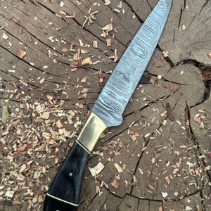 May include: A Damascus steel knife with a black handle and brass bolster. The blade has a distinctive pattern. The knife is shown on a wooden surface with wood shavings.