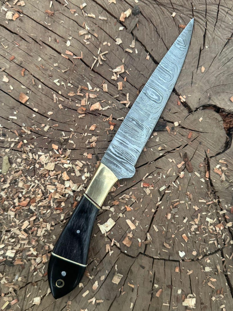 May include: A Damascus steel knife with a black handle and brass bolster. The blade has a distinctive pattern. The knife is shown on a wooden surface with wood shavings.
