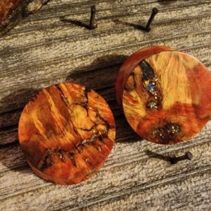 Könnte beinhalten: Zwei runde Holz-Ohrstecker mit einem natürlichen, rötlich-braunen Maserungsmuster. Die Stecker haben eine kreisförmige Form und sind wahrscheinlich für Ohrpiercings gedacht. Die Holzmaserung weist dunkle, komplizierte Muster und etwas schillernden Glitzer auf.
