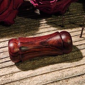 May include: A handcrafted, wooden bottle stopper with a rich, reddish-brown hue. The stopper features a bulbous top and a cylindrical body, showcasing the natural wood grain. The design is elegant and functional.