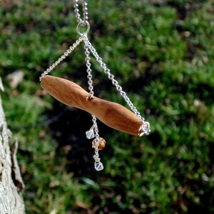 Könnte beinhalten: Eine silberne Kettenkette mit einem hölzernen Anhänger in Form eines kleinen Zweigs. Der Anhänger hängt an einer silbernen Kette mit kleinen klaren und bernsteinfarbenen Perlen.