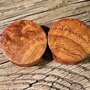 Puede incluir: Dos tapones redondos de madera, de color marrón intenso y con patrones de vetas únicos. Los tapones son circulares y parecen hechos a mano, mostrando la belleza natural de la madera. La veta de la madera crea un diseño orgánico y arremolinado.