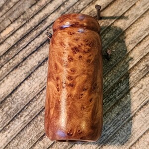 May include: A handcrafted wooden bottle stopper with a rich, brown burl wood grain pattern. The stopper has a rounded top and a cylindrical body. Two small metal pins are attached to the top. The stopper is displayed on a weathered wooden surface.