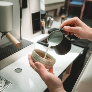 May include: A matte black stainless steel milk steaming pitcher pouring frothed milk into a small, round, beige and white marbled ceramic cup. The cup is held in one hand while the pitcher is held in the other, with an espresso machine visible nearby.