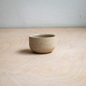 Peut inclure: Un petit bol rond en céramique avec une base de couleur beige clair et un motif moucheté composé de points brun foncé et noirs sur toute la surface extérieure. Le bol présente une silhouette lisse et incurvée avec une large ouverture.