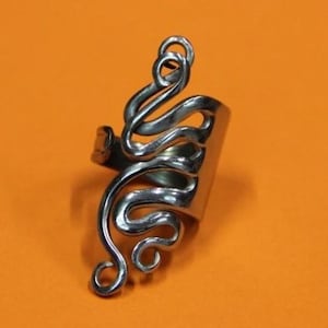 May include: A silver ring with a unique, abstract design. The ring is made of metal and has a wide band. The design is intricate and features a series of curves and loops.