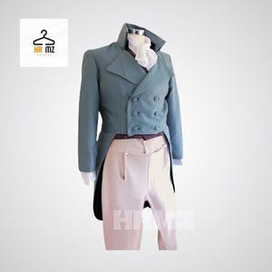Könnte beinhalten: Eine tealfarbene, zweireihige Jacke mit hohem Kragen und schwarzen Schößen, kombiniert mit cremefarbener Hose. Ein weißes Jabot ist am Hals zu sehen. Das Outfit wird auf einer Schaufensterpuppe präsentiert. Das HR MZ-Logo befindet sich oben links.
