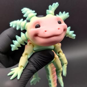 May include: A 3D printed articulated axolotl figure featuring a gradient colour scheme that transitions from soft pink on the face to light green and yellow along the body, limbs, and external gills, with two small black eyes and a smiling mouth.