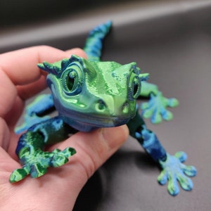 May include: A 3D printed green and blue gecko figurine with a textured surface. The gecko has large eyes and a wide mouth.