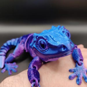 May include: A 3D printed articulated lizard figurine featuring a vibrant colour-shifting finish that transitions between shades of metallic blue and deep purple. The lizard has a textured surface, large eyes, and segmented limbs that allow for movement.
