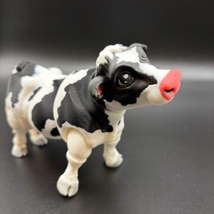 May include: A 3D-printed cow figurine with a black and white spotted pattern. The cow has a pink nose and detailed features. The figurine is standing on a dark surface, showcasing a farm animal theme.