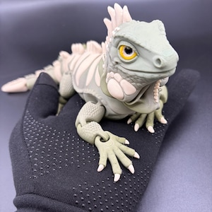 May include: An articulated iguana figurine featuring a sage green body with cream-coloured accents along the dorsal spines, limbs, and throat. The reptile has prominent yellow eyes with black pupils and a textured surface finish throughout its body.