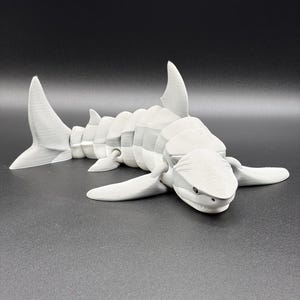 May include: A white, articulated shark toy with a segmented body, fins, and a detailed head. The toy is lying on a dark surface, showcasing its design and structure. The shark's jaws are slightly open, and the eyes are visible.