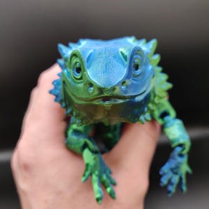 May include: A 3D-printed iguana figurine in shades of blue and green. The lizard is held in a hand, showcasing its detailed features, including textured skin and articulated limbs. The figurine has a gradient colour effect, transitioning from blue to green.