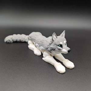 May include: A grey and white articulated wolf figurine in a crouching pose. The wolf has green eyes and a black nose. The tail is long and bushy. The figurine is on a dark grey surface.