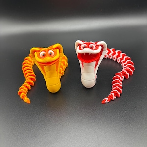 May include: Two 3D-printed toy snakes. One is yellow and orange, the other is red and white. Both have a cobra-like head and a segmented body. The snakes are posed on a dark surface.
