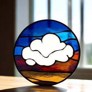 Op de afbeelding: Een rond stuk glas-in-lood dat een witte wolk tegen een zonsondergangachtergrond afbeeldt. De zonsondergang heeft tinten van blauw, oranje en rood.
