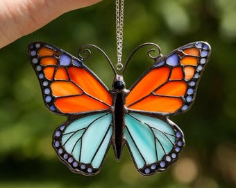May include: A stained glass butterfly ornament with orange and blue wings, outlined in black. The butterfly is suspended by a silver chain. The wings are divided into two sections, with the top half orange and the bottom half blue.
