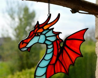 May include: Stained glass dragon suncatcher with a red, orange, and light blue color scheme. The dragon has a detailed head, a wing, and is suspended by a chain. The artwork is displayed in front of a blurred green background.