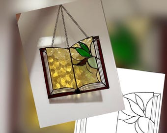 Puede incluir: Libro abierto de vidrieras con una cadena para colgar. El libro tiene un marco marrón oscuro, páginas de vidrio amarillo y verde y un diseño de hojas. Un dibujo lineal en blanco y negro del libro está en el fondo.