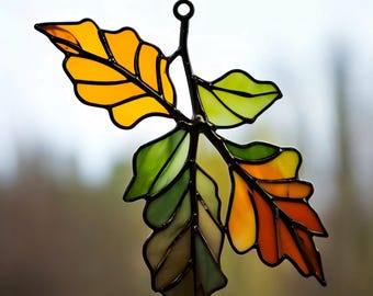 May include: Stained glass leaf decoration with a black metal frame. The leaves are in shades of yellow, orange, and green, with a dark brown stem and a hanging chain. The leaf design is reminiscent of autumn.