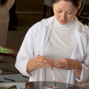 May include: A woman in a white lab coat and turtleneck is working on jewelry. Several copper and silver bracelets are on a reflective surface. The woman is holding a copper wire, possibly creating a bracelet.