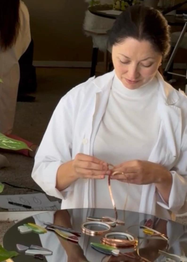 May include: A woman in a white lab coat and turtleneck is working on jewelry. Several copper and silver bracelets are on a reflective surface. The woman is holding a copper wire, possibly creating a bracelet.