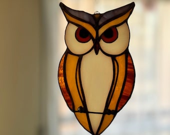 May include: A stained glass owl suncatcher with a black outline. The owl has a cream-colored body, orange wings, and red eyes. The top of the owl's head is dark purple with yellow accents. A small black wire is attached for hanging.
