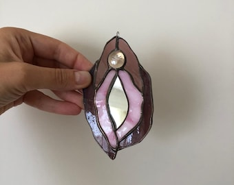 May include: Stained glass art piece in the shape of a stylized vulva. The piece features shades of pink and brown glass, with a mirrored center and a small, clear gem at the top. The edges are outlined in a dark metal.