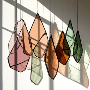 Puede incluir: Una colección de paneles de vidrieras colgantes en varias formas y colores. Los paneles son en tonos de rosa, melocotón, naranja y verde, con marcos de metal negro. La luz del sol proyecta sombras en la pared blanca detrás de ellos.